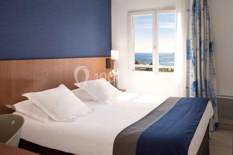 Chambre lumineuse avec lit double, décor bleu et blanc, fenêtre offrant une vue sur la mer.