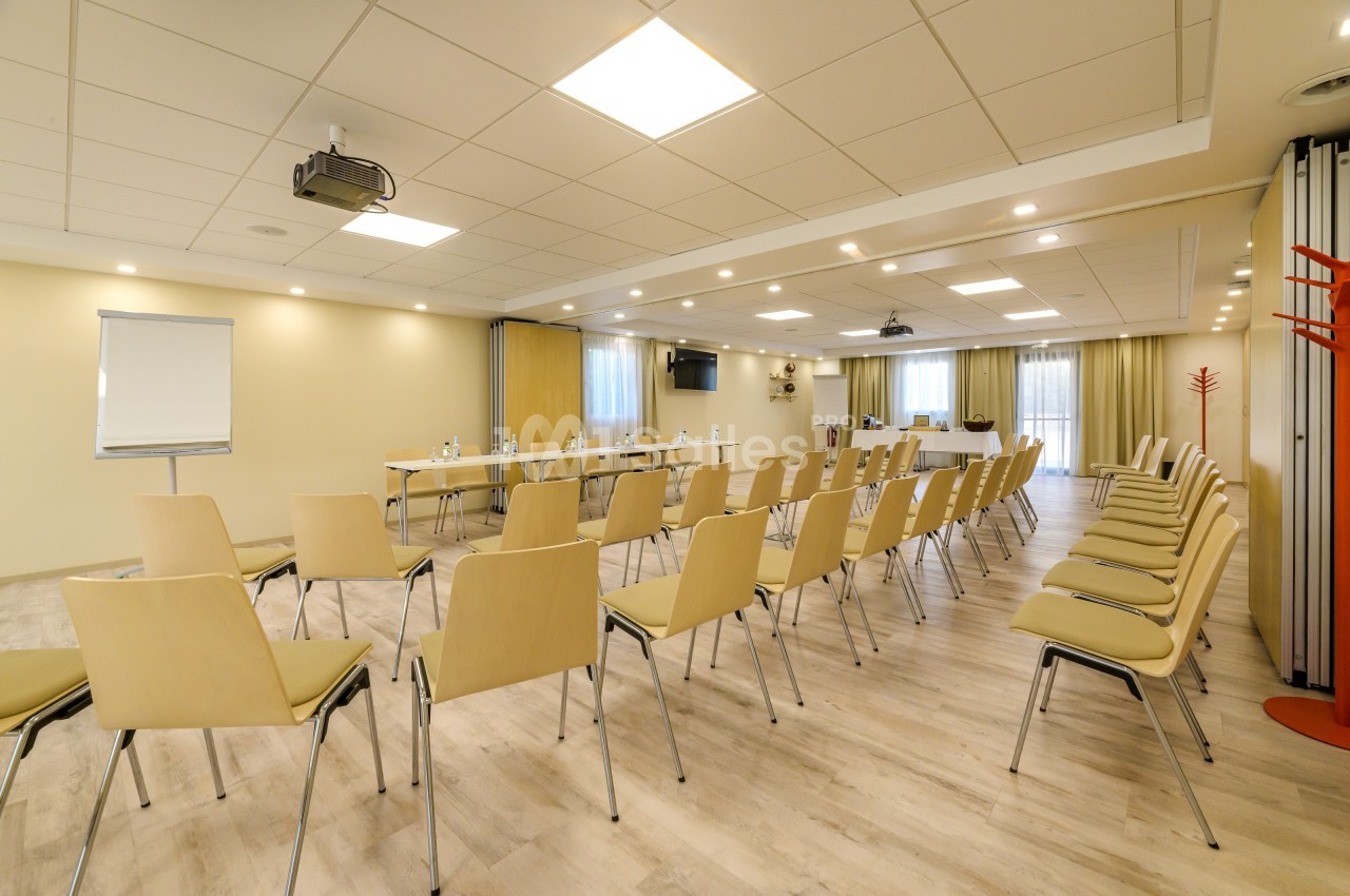 Salle de conférence lumineuse avec des rangées de chaises en bois, un tableau blanc et un projecteur au plafond.