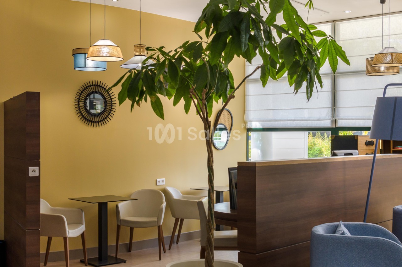 Espace intérieur lumineux avec des tables, des chaises, un grand arbre décoratif et des luminaires suspendus.