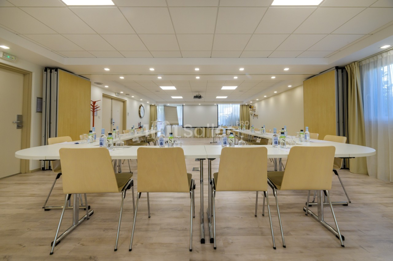 Salle de réunion lumineuse avec tables disposées en U, chaises en bois clair et écran de projection au fond.