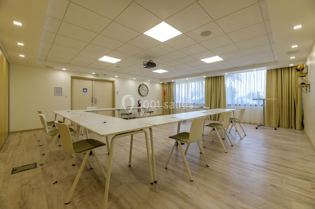Salle de réunion lumineuse avec tables blanches disposées en U, chaises beige et grand écran au plafond.