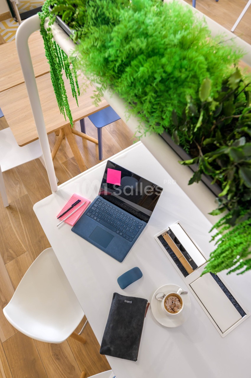 Ordinateur portable ouvert sur une table blanche avec carnet, tasse de café et plantes suspendues au-dessus.