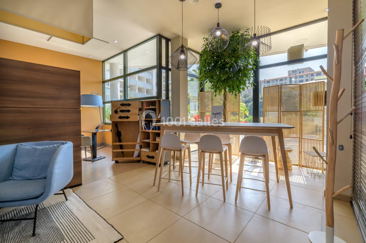 Espace lumineux avec table haute, tabourets, plantes suspendues et grande baie vitrée donnant sur une terrasse.