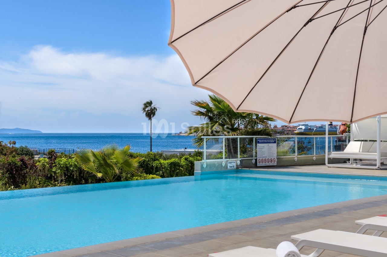 Piscine extérieure avec vue sur la mer, entourée de palmiers et protégée par un grand parasol beige.