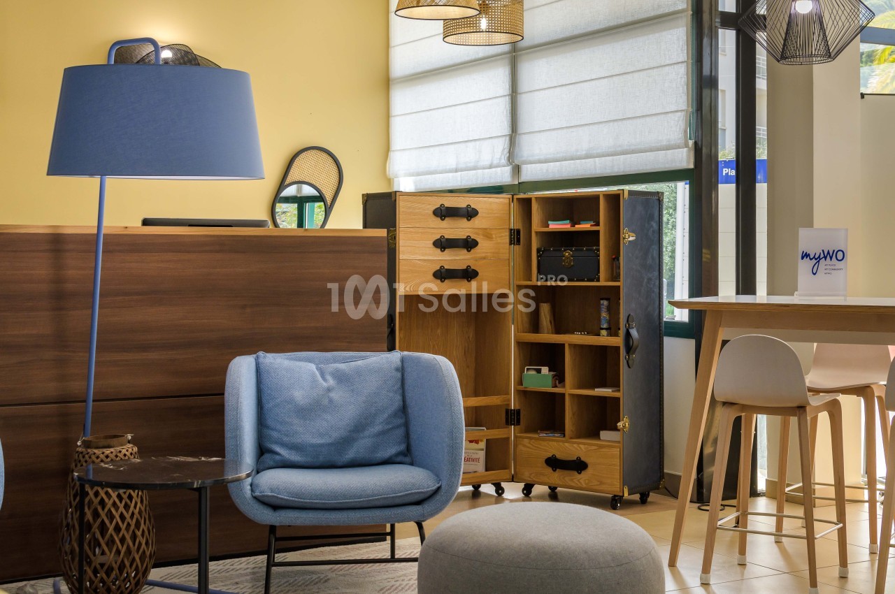 Espace intérieur lumineux avec fauteuil bleu, lampadaire, meuble ouvert en bois et table haute près d'une fenêtre.