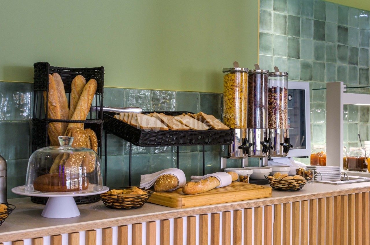 Buffet de petit-déjeuner avec pains, céréales, confitures et viennoiseries sur un comptoir en bois devant un mur vert.