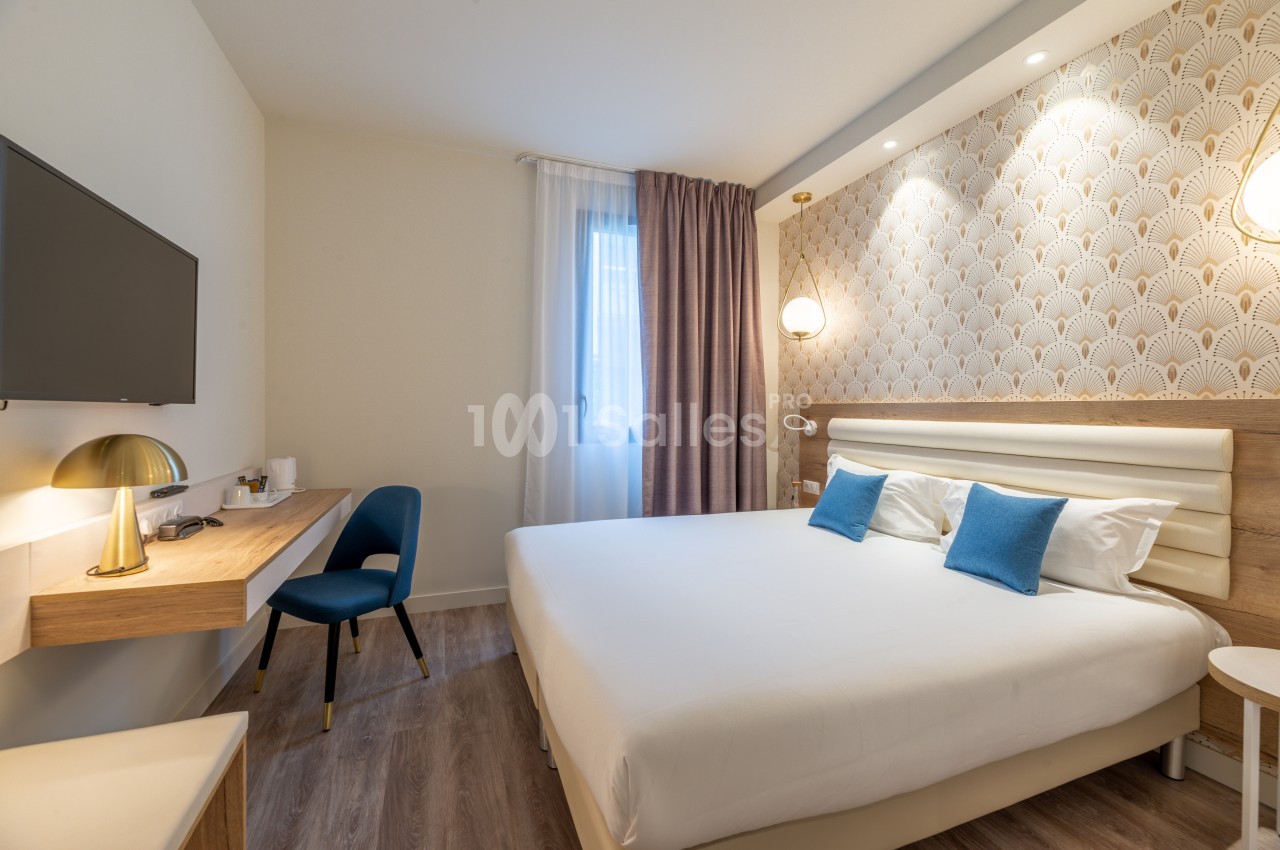 Chambre d'hôtel lumineuse avec un lit double, deux coussins bleus, un bureau, une chaise et une télévision murale.