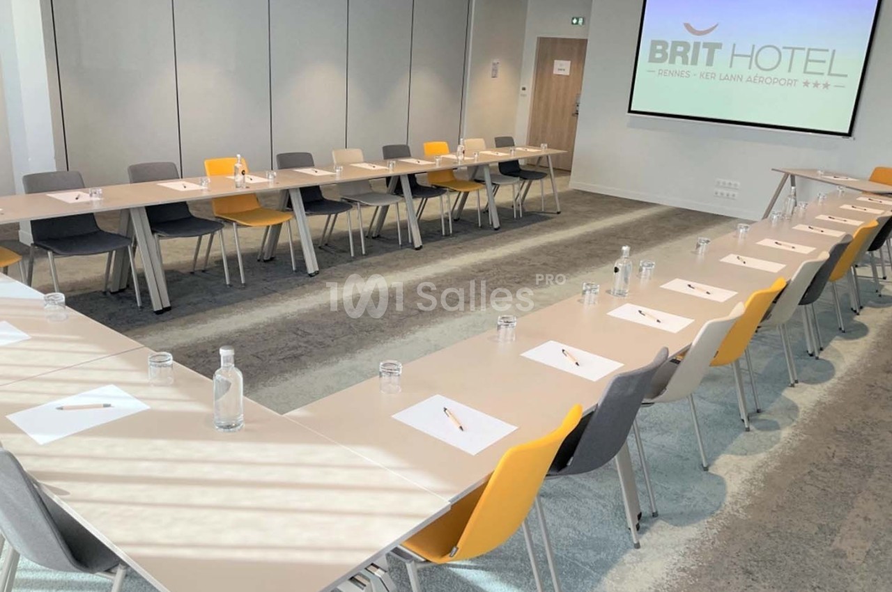 Salle de réunion lumineuse avec tables disposées en U, chaises colorées et fournitures de bureau sur chaque place.