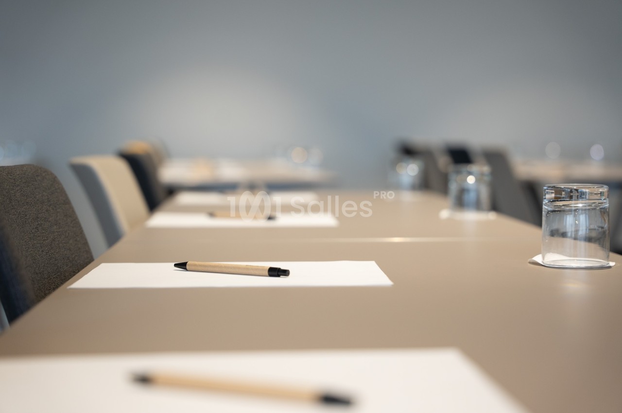 Des tables alignées avec des feuilles blanches, des stylos et des verres d'eau dans une salle de réunion vide.