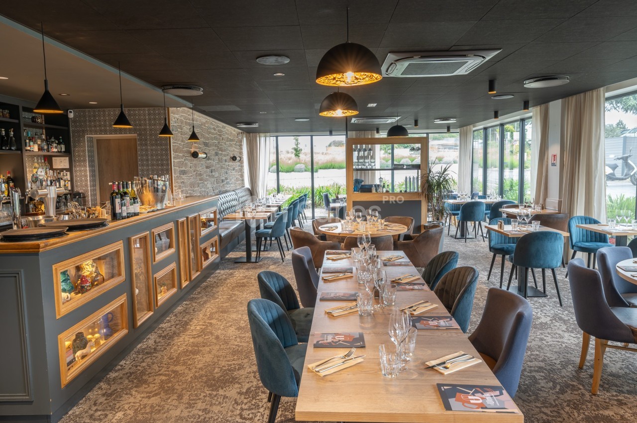 Salle de restaurant moderne avec tables dressées, chaises rembourrées, bar éclairé et grandes baies vitrées donnant sur l…