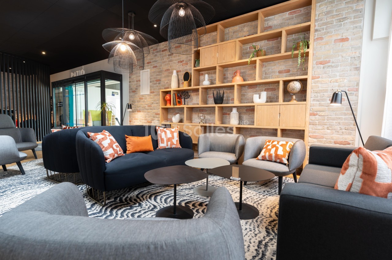 Espace salon moderne avec canapés, fauteuils, tables basses et étagères décoratives sur un mur en briques.