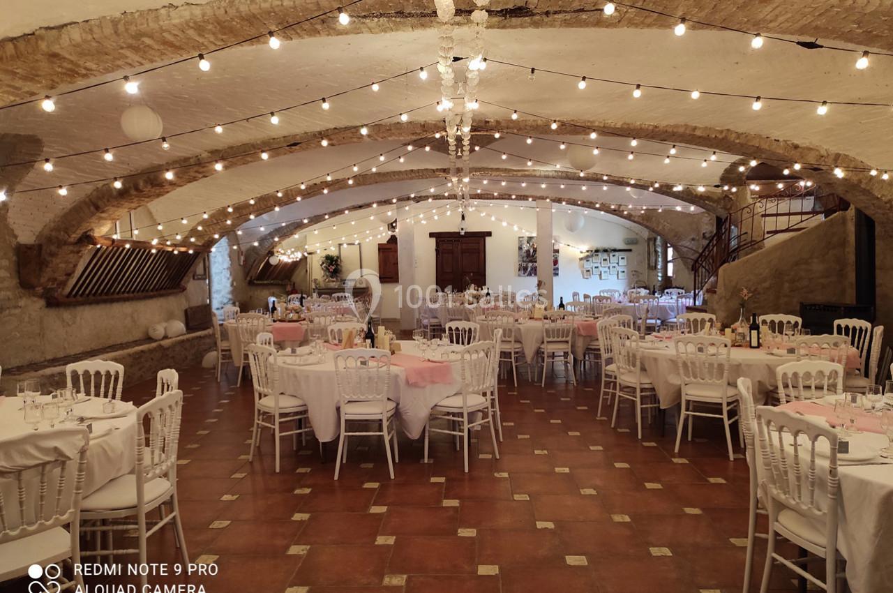 Salle voûtée décorée de guirlandes lumineuses, tables rondes dressées avec nappes blanches et chaises élégantes.