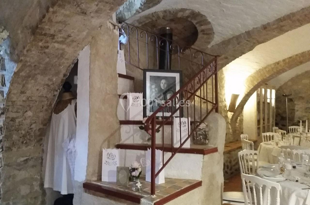 Escalier en pierre avec rampe en fer forgé dans une salle voûtée, décorée pour un événement avec des tables dressées.