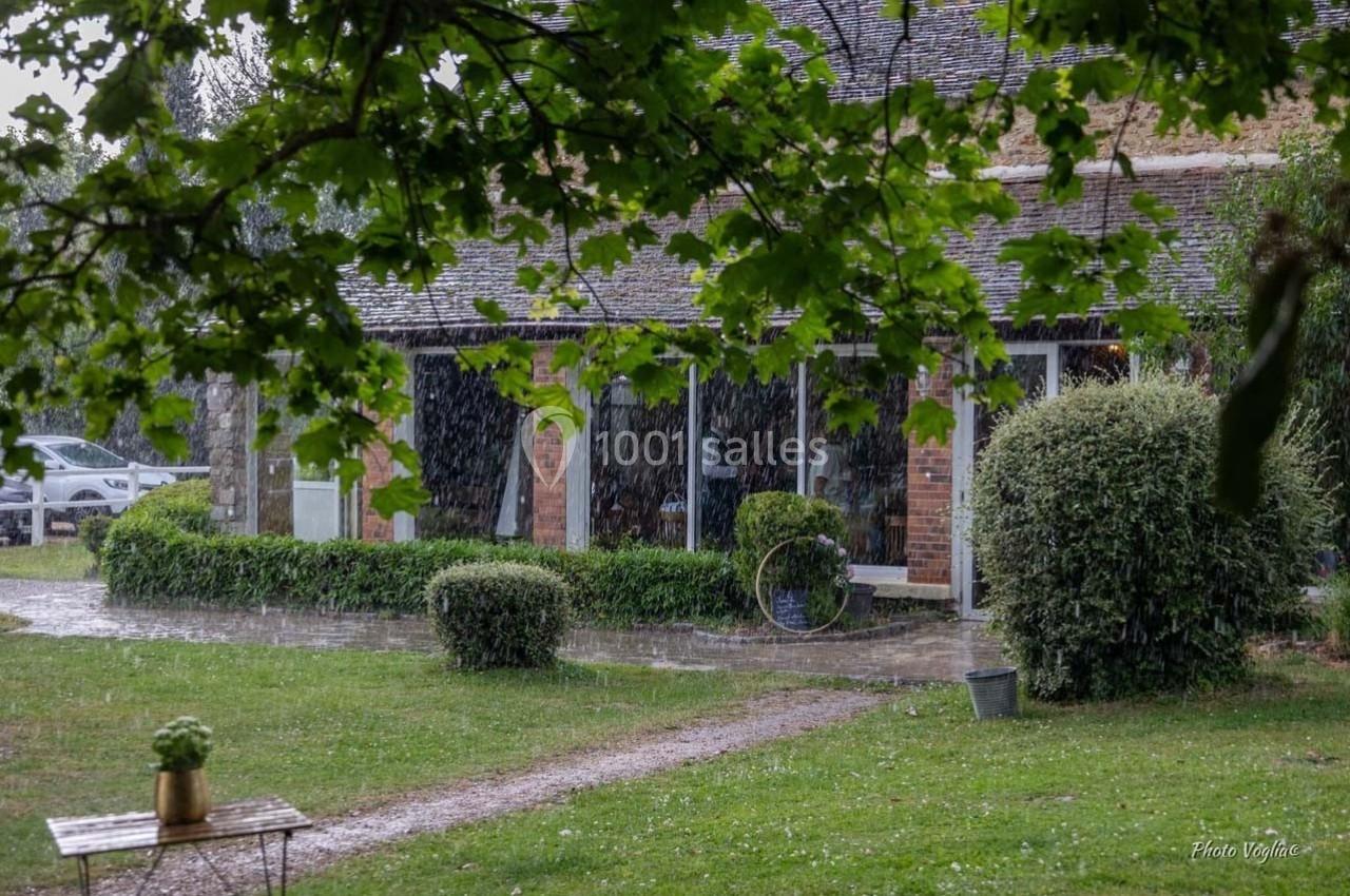 Maison en briques entourée de verdure sous une pluie fine, avec des arbres et un chemin en premier plan.