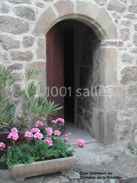 Porte en pierre ouverte sur un mur ancien, entourée de fleurs roses dans des jardinières en pierre.