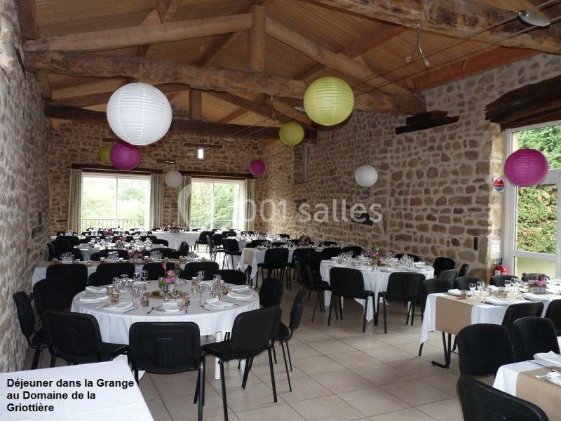 Salle en pierre avec tables rondes dressées pour un repas, décorée de lanternes suspendues et baignée de lumière naturelle.