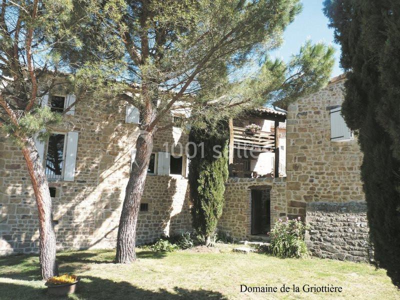 Façade en pierre d'une maison entourée d'arbres, avec des volets blancs et un espace extérieur ombragé.
