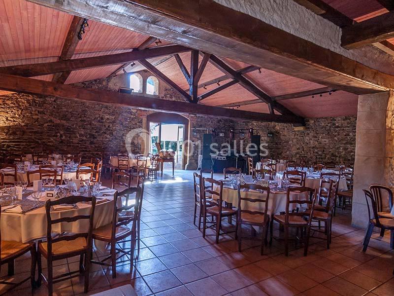 Salle de réception en pierre avec poutres apparentes, tables rondes dressées et lumière naturelle entrant par les fenêtres.