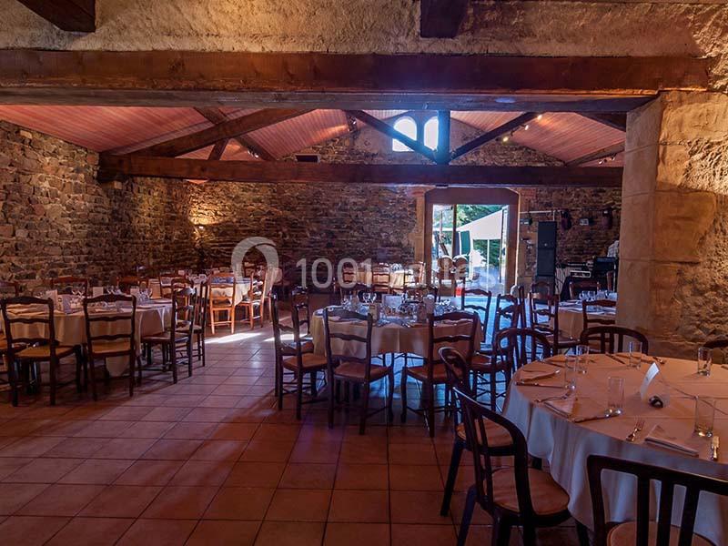 Salle en pierre avec poutres apparentes, tables rondes dressées pour un repas, lumière naturelle entrant par une porte…