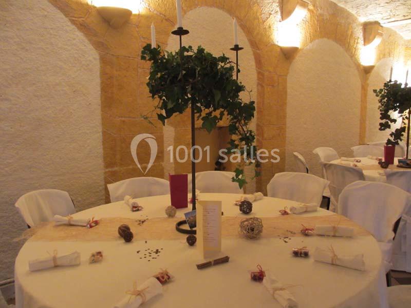 Salle décorée pour un événement, avec tables rondes dressées, nappes blanches et centres de table ornés de feuillage.