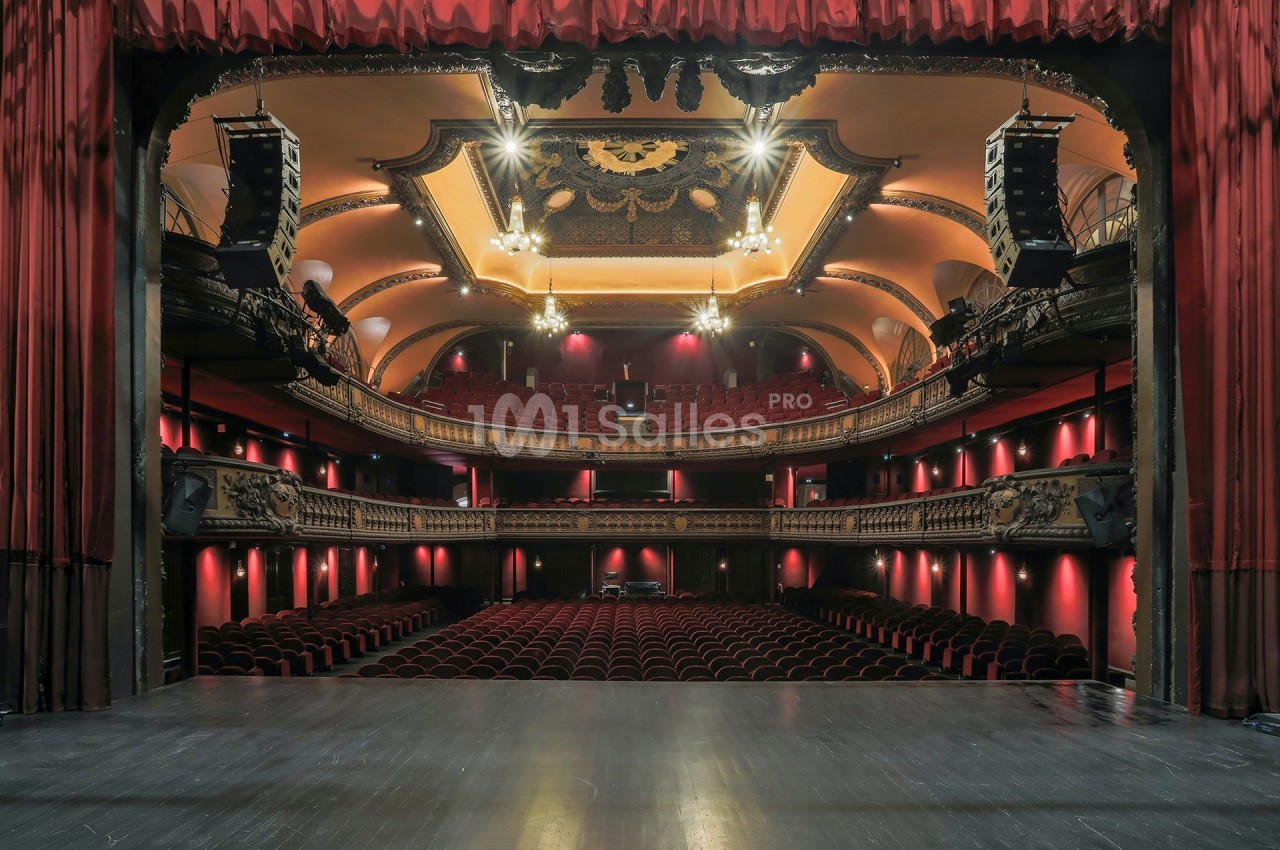 Vue depuis la scène d'un théâtre avec balcons ornés, lustres suspendus et rangées de sièges rouges vides.