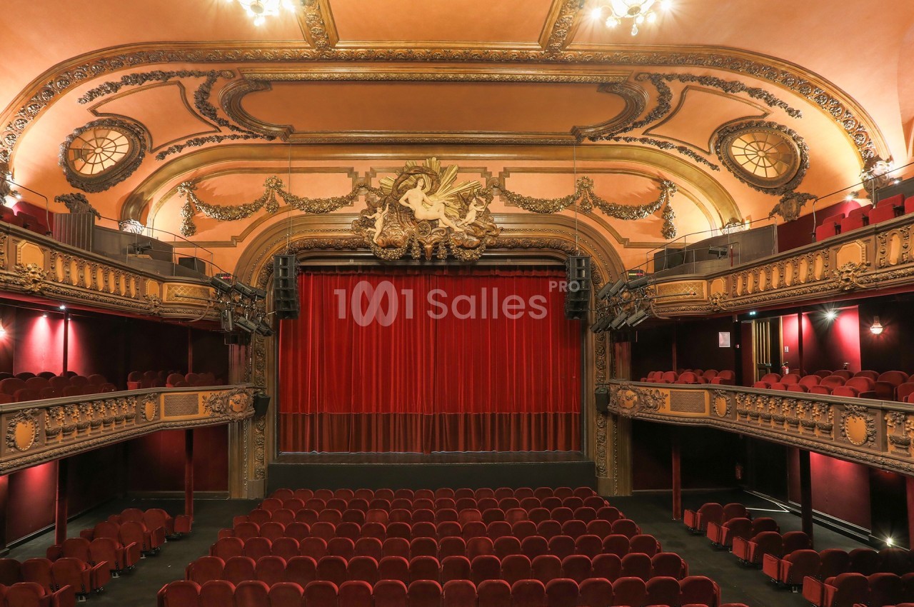 Intérieur d'un théâtre avec des sièges rouges, une scène encadrée de rideaux et une décoration ornée au plafond.