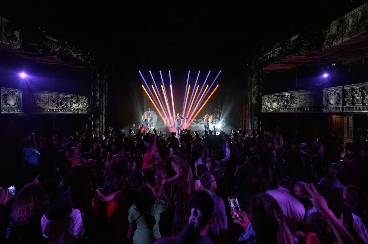Salle de concert avec un public nombreux et un groupe sur scène éclairé par des projecteurs colorés.