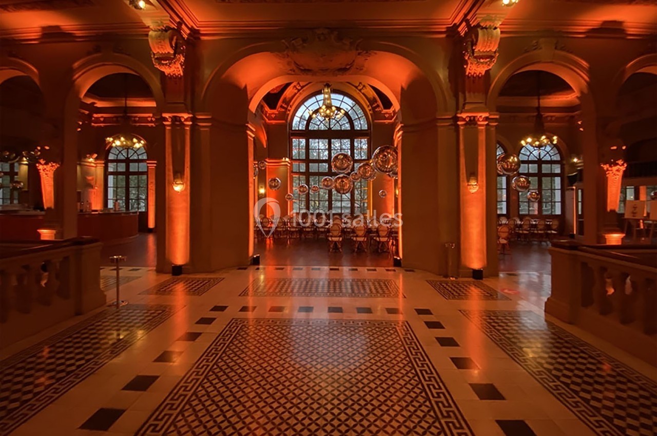 Salle élégante avec arches, sols carrelés et éclairage orange, tables alignées devant de grandes fenêtres.