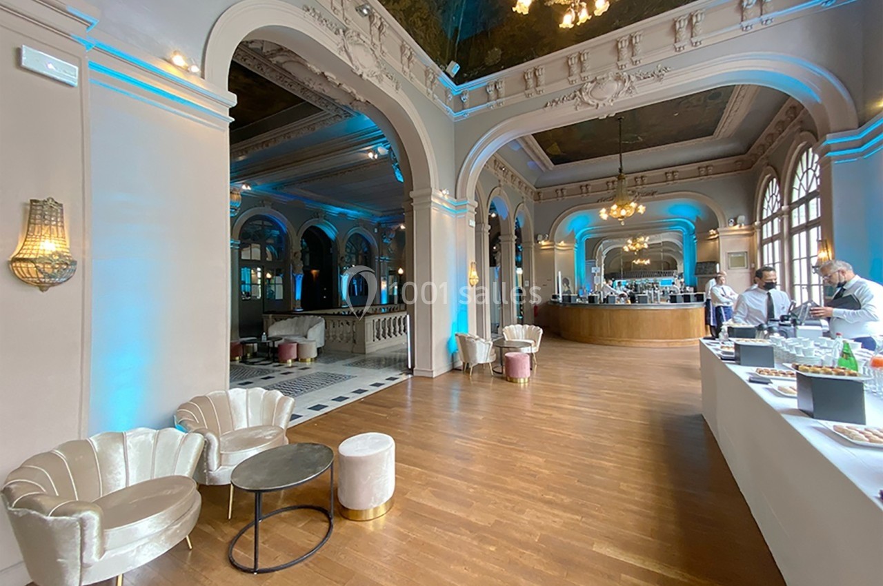 Salle élégante avec parquet, fauteuils en velours, arches décoratives et un espace de restauration en arrière-plan.