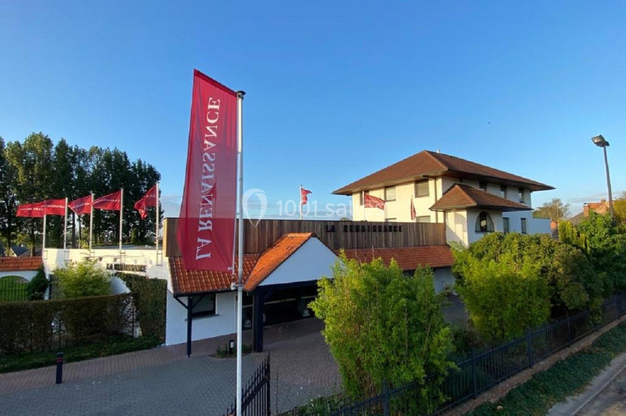Bâtiment moderne avec toit en tuiles rouges, entouré de végétation, et drapeaux rouges portant l'inscription ’Renaissance’.