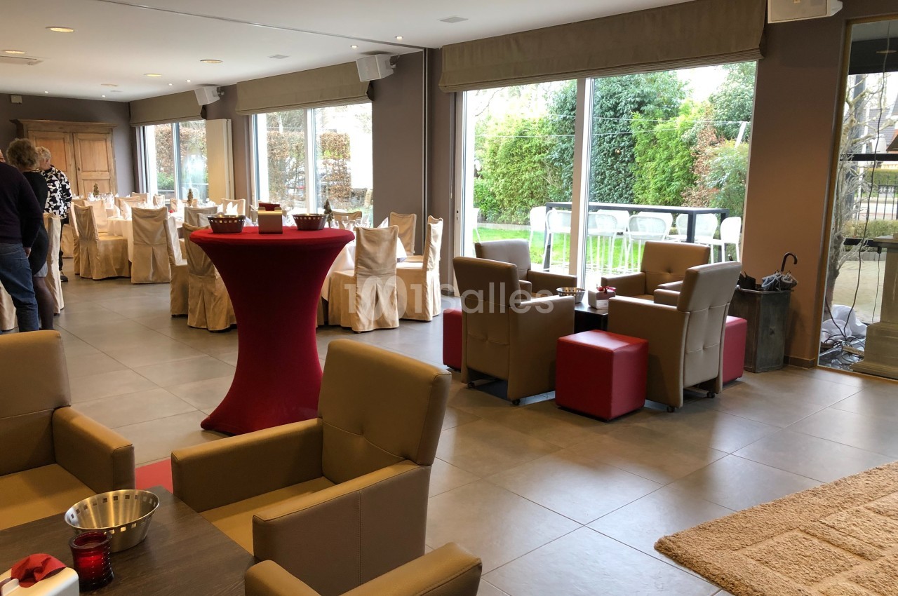 Salle lumineuse avec grandes baies vitrées, tables dressées, fauteuils beiges et tables hautes rouges.