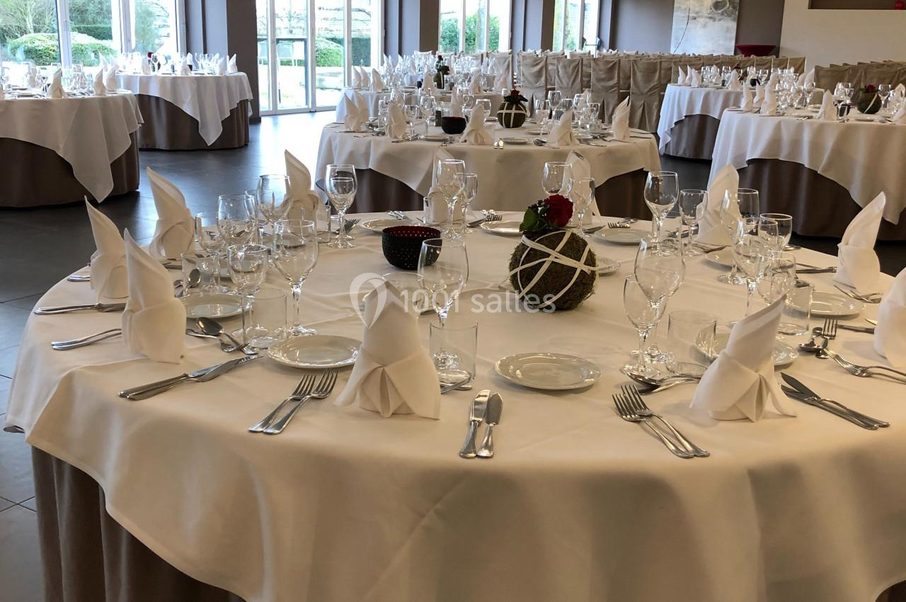 Salle de réception avec tables rondes dressées, nappes blanches, serviettes pliées et vaisselle élégante.