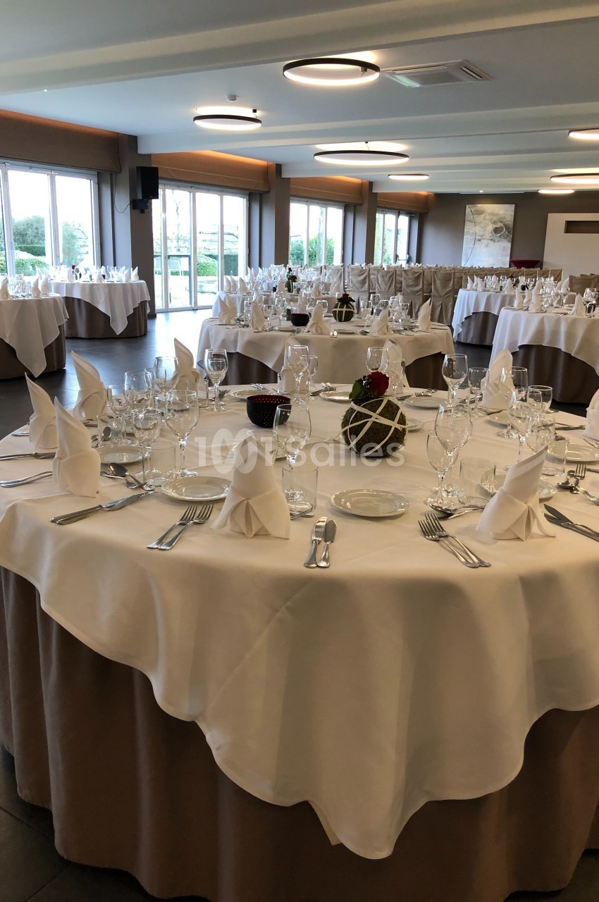 Salle de réception avec tables rondes dressées, nappes blanches, serviettes pliées et vaisselle élégante.