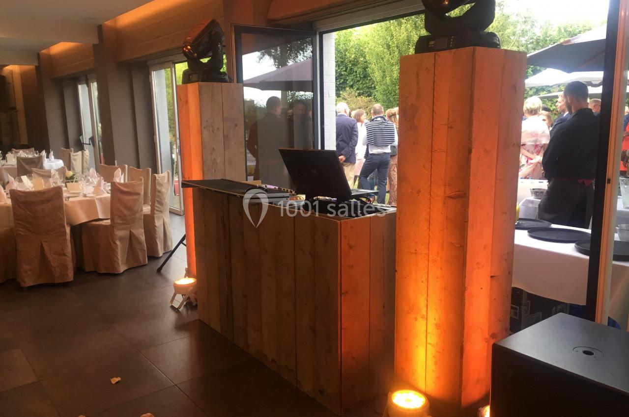 Console de DJ en bois avec équipement audio, éclairée par des spots, dans une salle avec vue sur un événement extérieur.