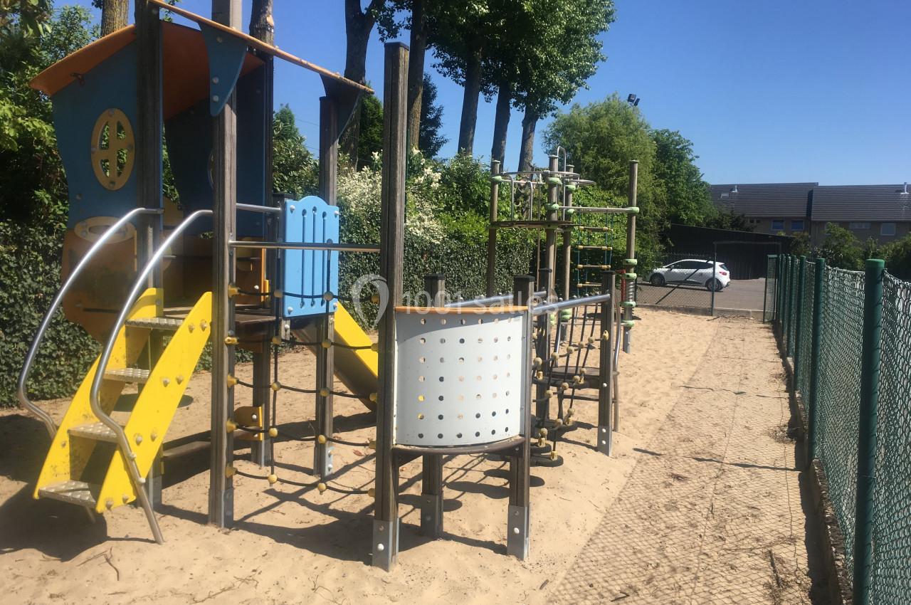 Structure de jeux en plein air avec toboggan et modules d'escalade, installée sur un sol sablonneux près d'une clôture.