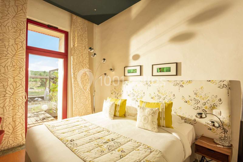 Chambre lumineuse avec lit double, décor floral, grandes fenêtres et vue sur un jardin ensoleillé.