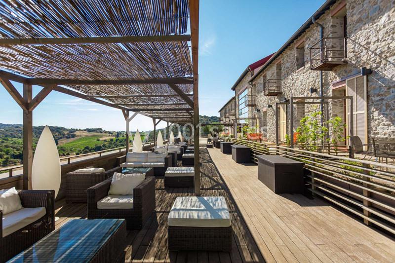 Terrasse en bois avec mobilier en rotin, pergola ombragée et vue sur une campagne vallonnée sous un ciel dégagé.