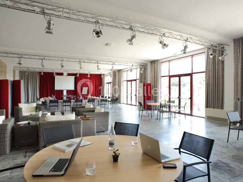 Salle lumineuse avec grandes baies vitrées, équipée de tables, chaises, canapés, et matériel de présentation.