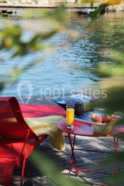 Fauteuil rouge et table avec jus d'orange et fruits, au bord d'un plan d'eau entouré de végétation.