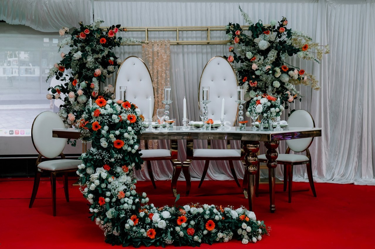 Table décorée pour un événement, entourée de chaises blanches et ornée de fleurs rouges, blanches et vertes.