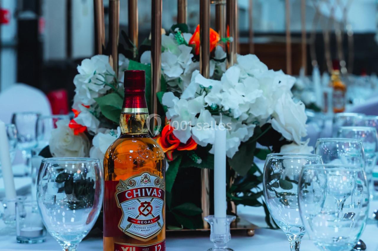 Bouteille de whisky posée sur une table décorée de fleurs blanches et rouges, entourée de verres et bougies.
