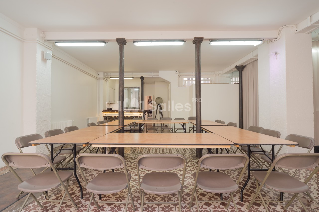 Salle lumineuse avec tables en bois disposées en rectangle, entourées de chaises pliantes, sur un sol en carreaux décoratifs.