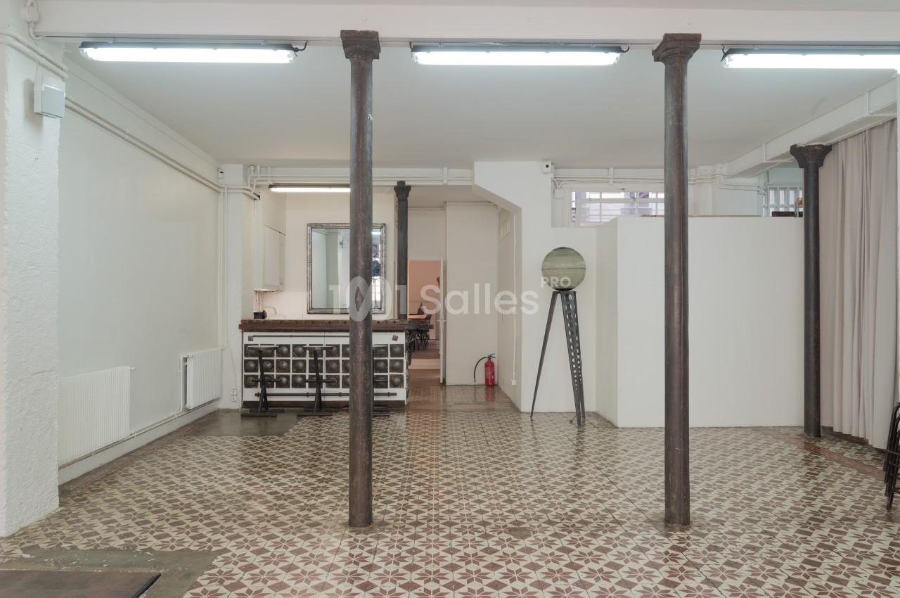 Intérieur d'un espace avec sol en carreaux de ciment, colonnes métalliques et vue sur une cuisine ouverte au fond.