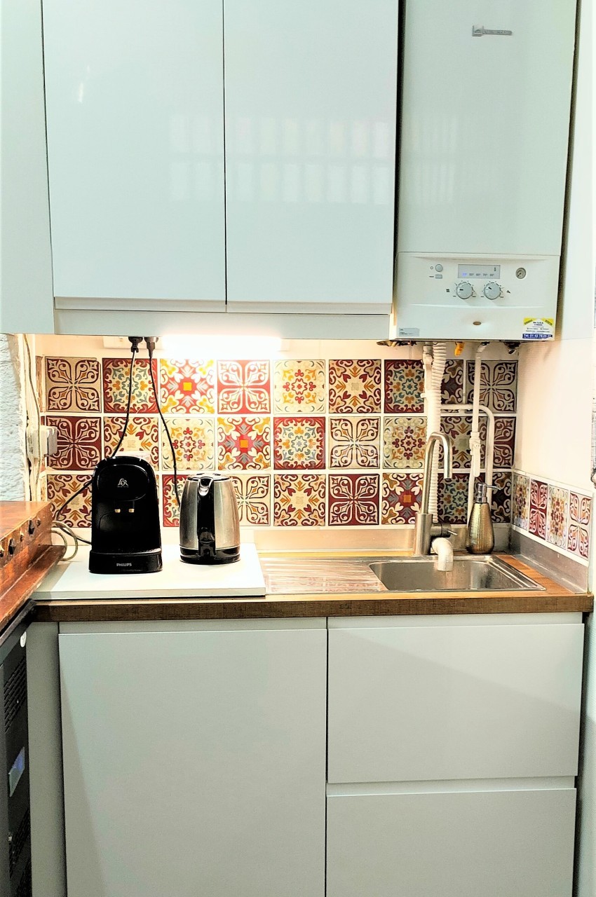 Cuisine compacte avec évier, robinet, carrelage coloré, chauffe-eau mural, cafetière et grille-pain sur le plan de travail.