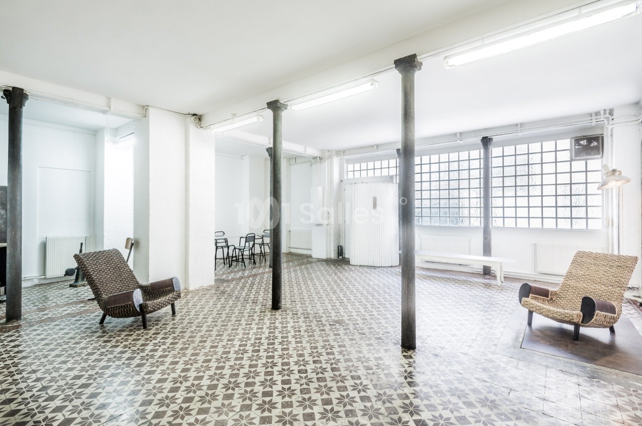 Salle lumineuse avec carrelage à motifs, colonnes métalliques, chaises et fauteuils en osier, grandes fenêtres en verre.