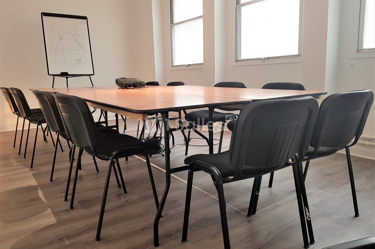 Salle de réunion avec une grande table en bois entourée de chaises noires, un tableau blanc et un projecteur.