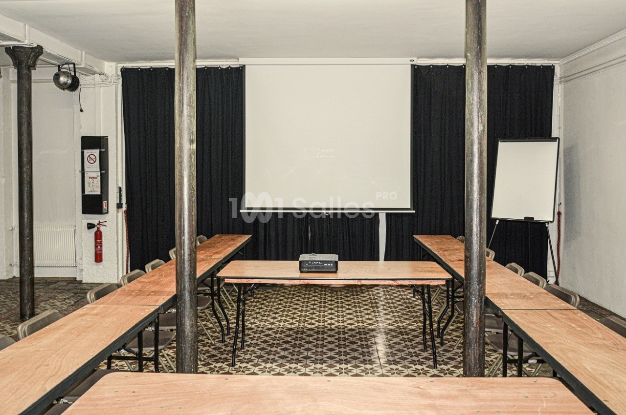 Salle de réunion avec tables en U, vidéoprojecteur, écran blanc et tableau de présentation.