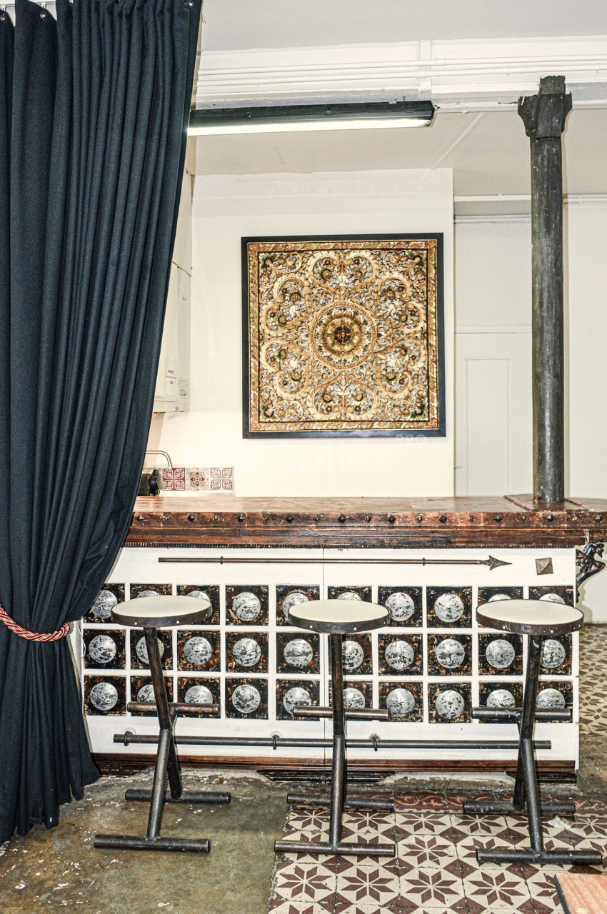 Comptoir en bois décoré, trois tabourets hauts, rideau noir à gauche et tableau ornemental au mur.