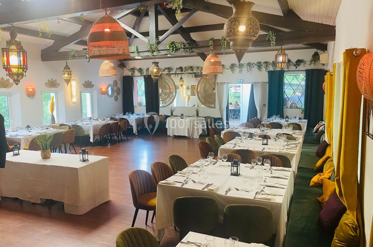 Salle de restaurant lumineuse avec tables dressées, chaises colorées, poutres apparentes et décorations suspendues.