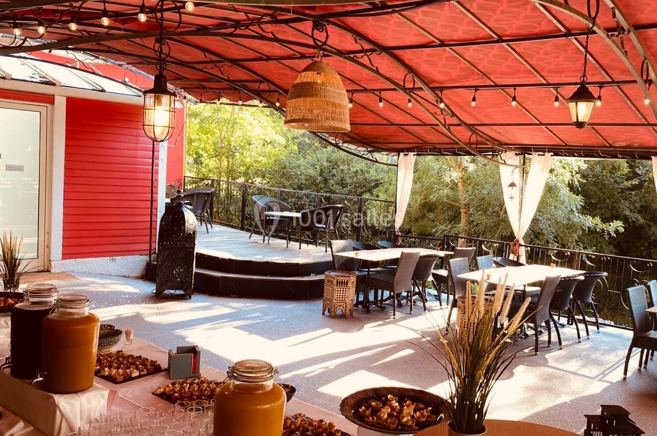 Terrasse couverte avec tables dressées, boissons et apéritifs disposés, entourée de verdure et éclairée par des guirlandes.